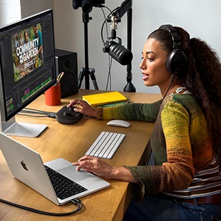 امرأة في منزلها تعمل خلف مكتبها على MacBook&nbsp;Pro متصل بشاشة خارجية وتستخدم سماعات رأس وتتحدث في الميكروفون