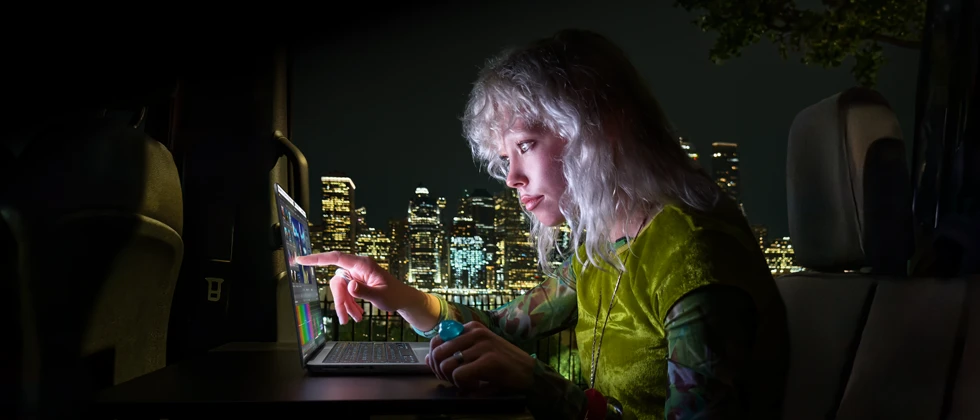 شخص يجلس في المقعد الخلفي لسيارة فان، باب الراكب المفتوح يكشف عن أفق مدينة في الليل، جهاز MacBook Pro مع شاشة PROLED XDR موضوع على طاولة عمل، ضوء الشاشة ينعكس على الشخص أثناء استخدامه لميزة اللمس من أجل تعديل فيديو في برنامج DaVinci Resolve Studio‏