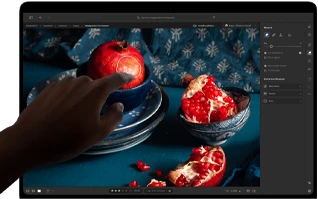 جهاز MacBook Pro مع شاشة PROLED XDR يعرض صورة ثابتة لثمار الرمان وقطع خزفية ومنسوجات زرقاء، مع يد تمتد نحو الشاشة للتفاعل مع العمل الفني