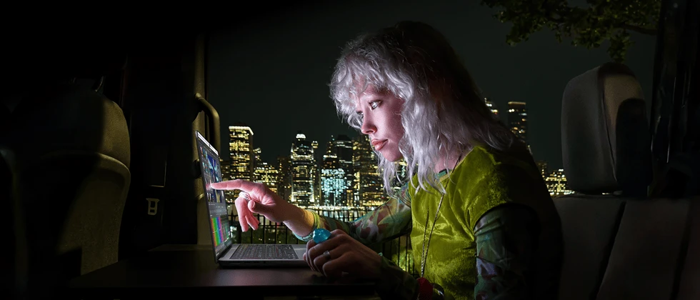A person sitting in the back seat of a van, the open passenger door reveals a city skyline at night, MacBook Pro with PROLED XDR Display on a worktable, the person is illuminated by the screen while using the Touch feature to edit a video in DaVinci Resolve Studio