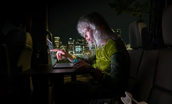 A person sitting in the back seat of a van, the open passenger door reveals a city skyline at night, MacBook Pro with PROLED XDR Display on a worktable, the person is illuminated by the screen while using the Touch feature to edit a video in DaVinci Resolve Studio