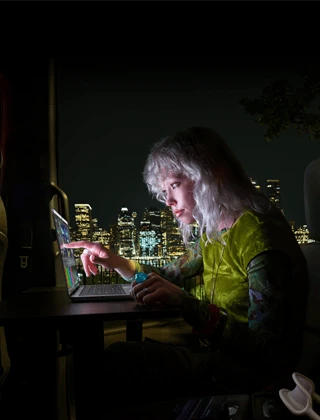 A person sitting in the back seat of a van, the open passenger door reveals a city skyline at night, MacBook Pro with PROLED XDR Display on a worktable, the person is illuminated by the screen while using the Touch feature to edit a video in DaVinci Resolve Studio